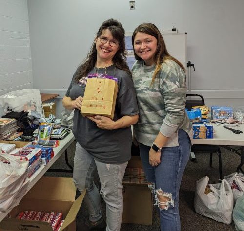Two women stand holding a brown bag filled with donations for seniors.