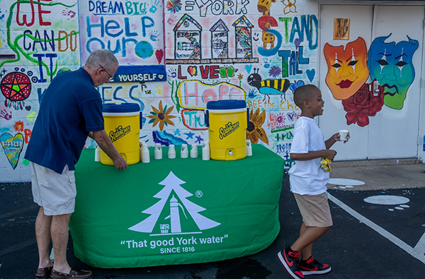 A man gets water from a water cooler that is on top of a York Water Company branded table. A boy who has just filled a cup of water from the table walks away.