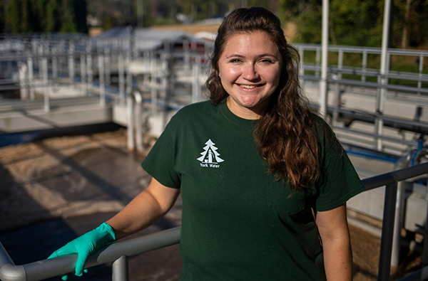 A woman smiles in the foreground. She is wearing a dark green York Water branded shirt. In the background is a wastewater treatment plant.