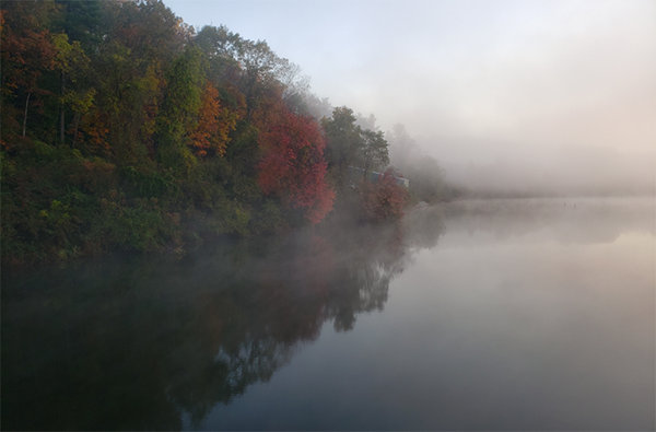 An early morning photograph of Lake Redman. Mist rises from the water. Trees on the shoreline show signs of fall. They are reflected in the water.